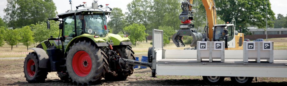 Photo of the practical test, on the left the automated tractor, on the right a trailer, behind the trailer an automated excavator loading hazardous goods